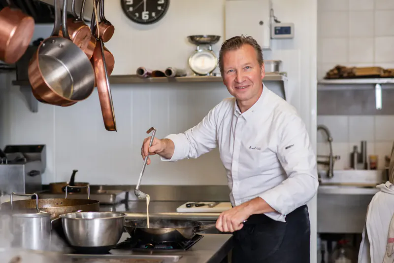 Smiling chef in white uniform stirring batter in pan with spatula in professional kitchen with copper pots