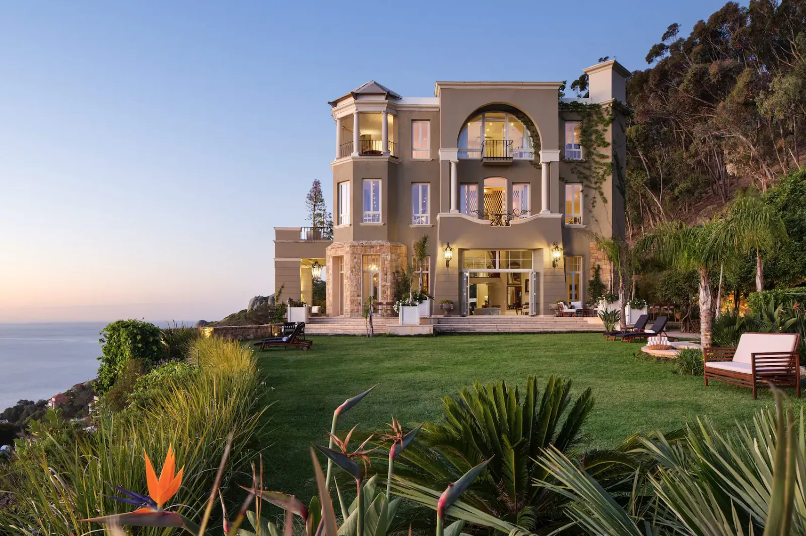 Luxurious beige mansion with arched windows and tower on green lawn overlooking Atlantic Ocean at sunset, Cape Town.