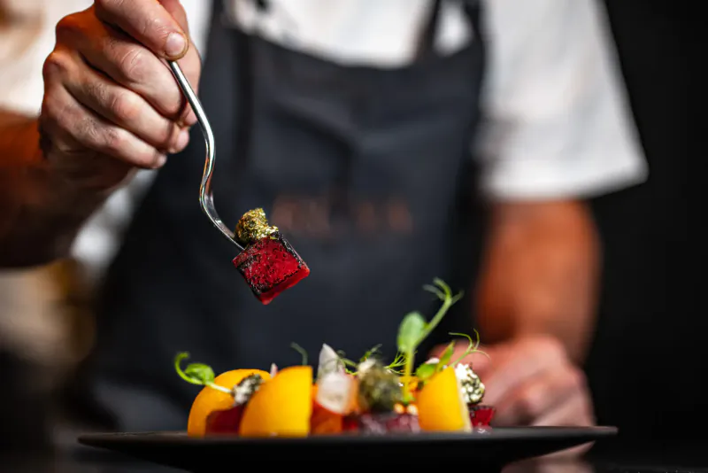 Chef's hand in black apron forking red beet on plate with peas, apricots, and garnishes