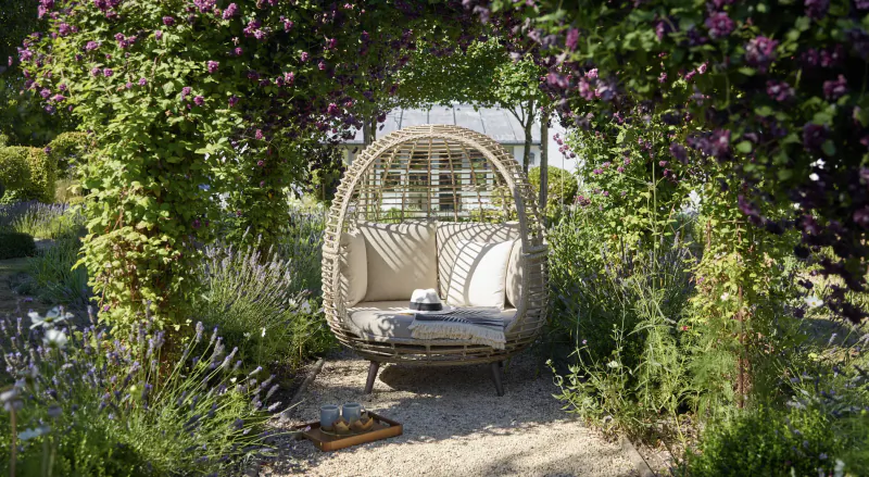White egg-shaped hanging chair with cushions and hat on tray, framed by lush purple bougainvillea arch in garden.