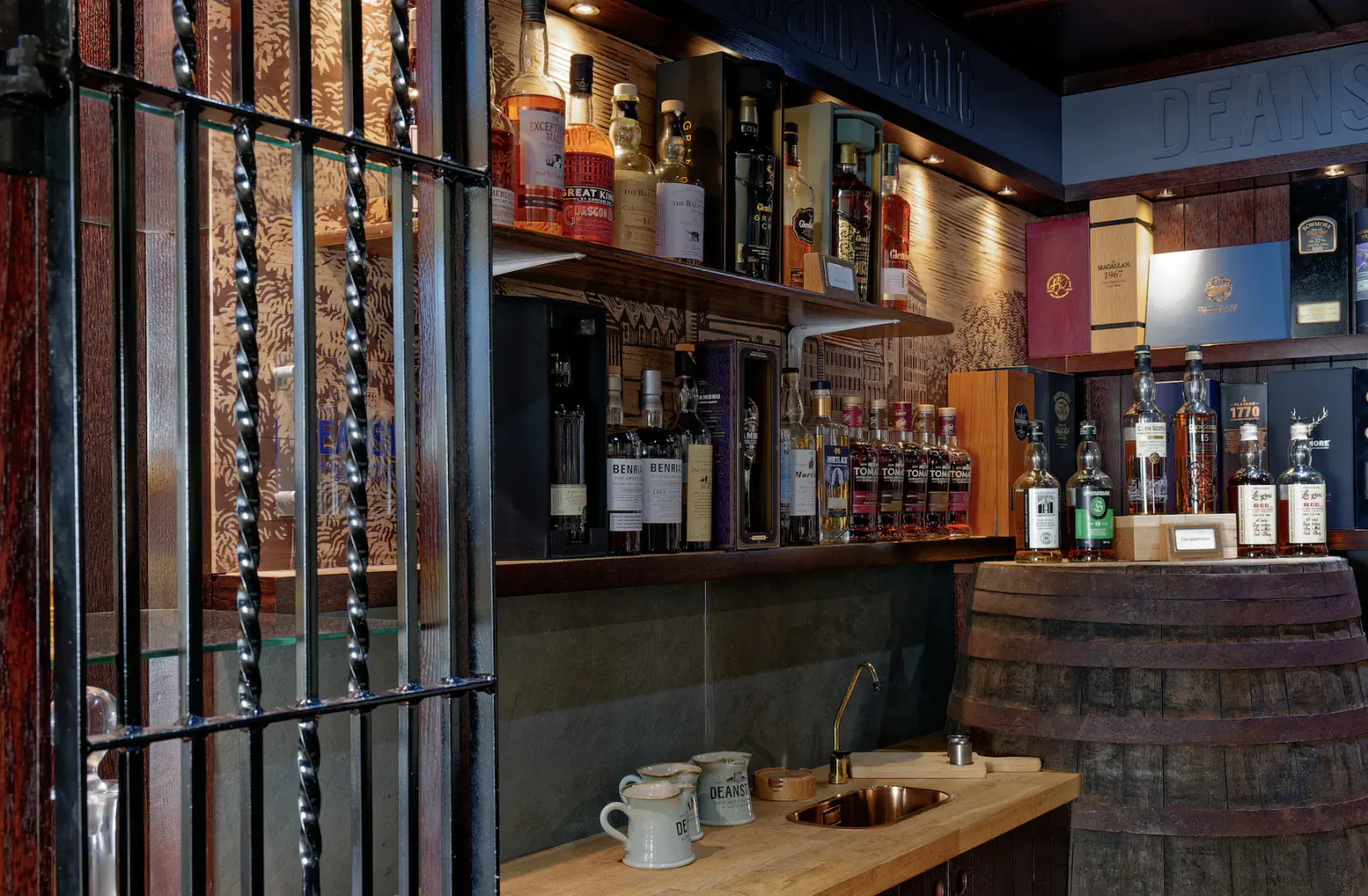 Curated whiskey selection on shelves behind iron bars in dimly lit bar, with wooden barrel and counter.