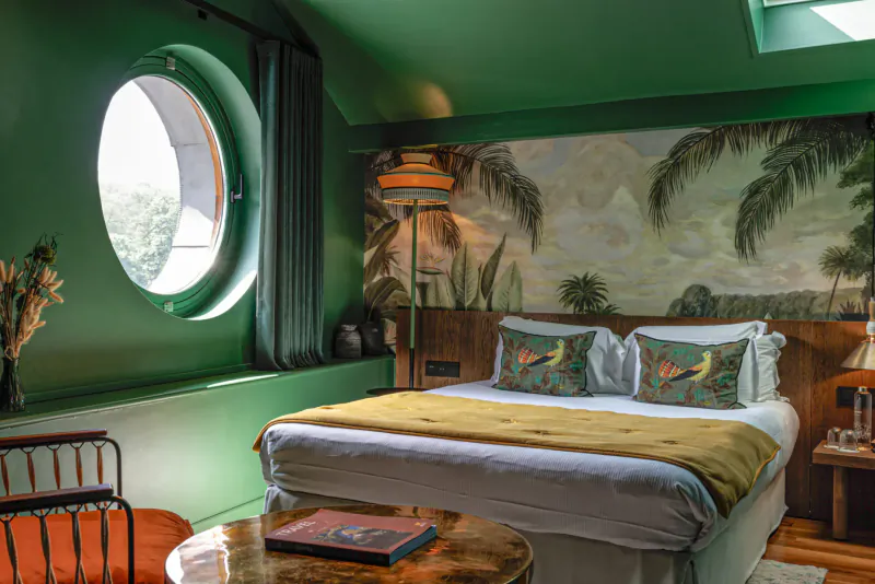 Cozy green attic hotel room with porthole window, palm mural, yellow bed, and rattan chair at Les Étangs de Corot Paris