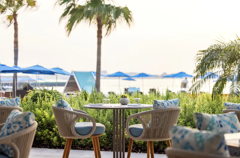 Outdoor dining table with blue cushions at Four Seasons Resort Dubai, surrounded by palms, blue umbrellas, and sea view