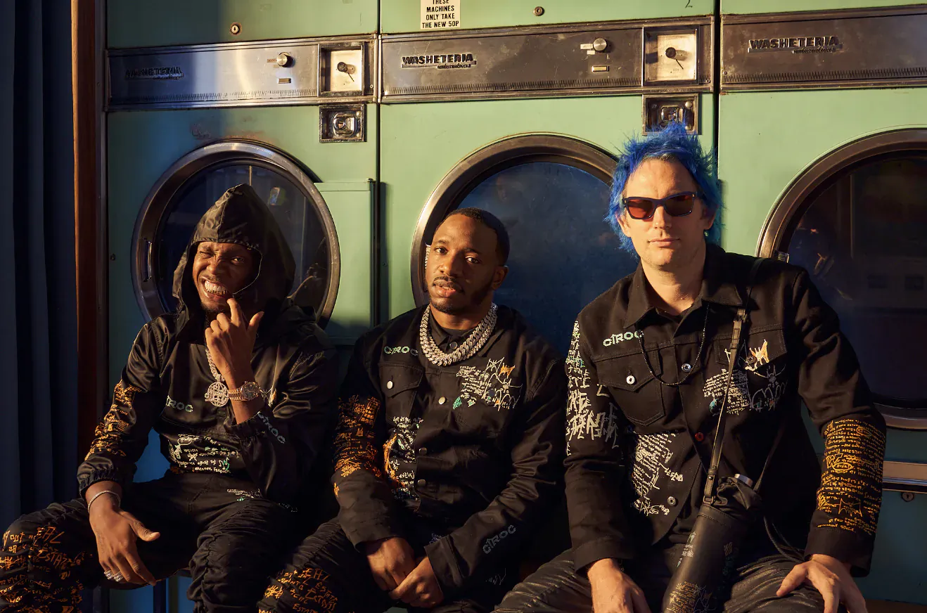 Krept & Konan with MJB posing coolly in front of green laundry machines, wearing stylish black outfits with gold chains.