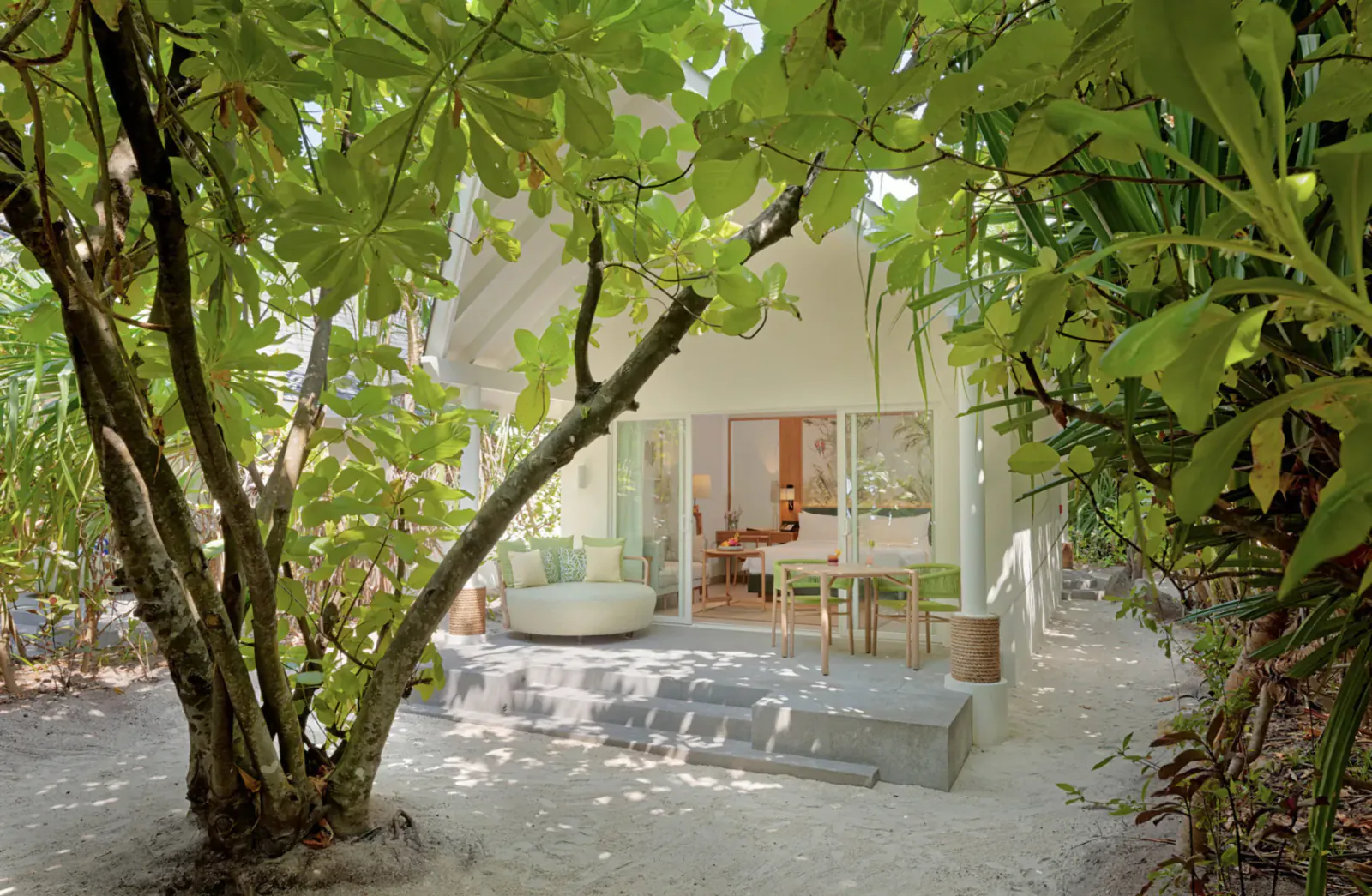 Amari Raaya Maldives luxury overwater villa with open glass doors, lush green trees, outdoor seating on sandy deck