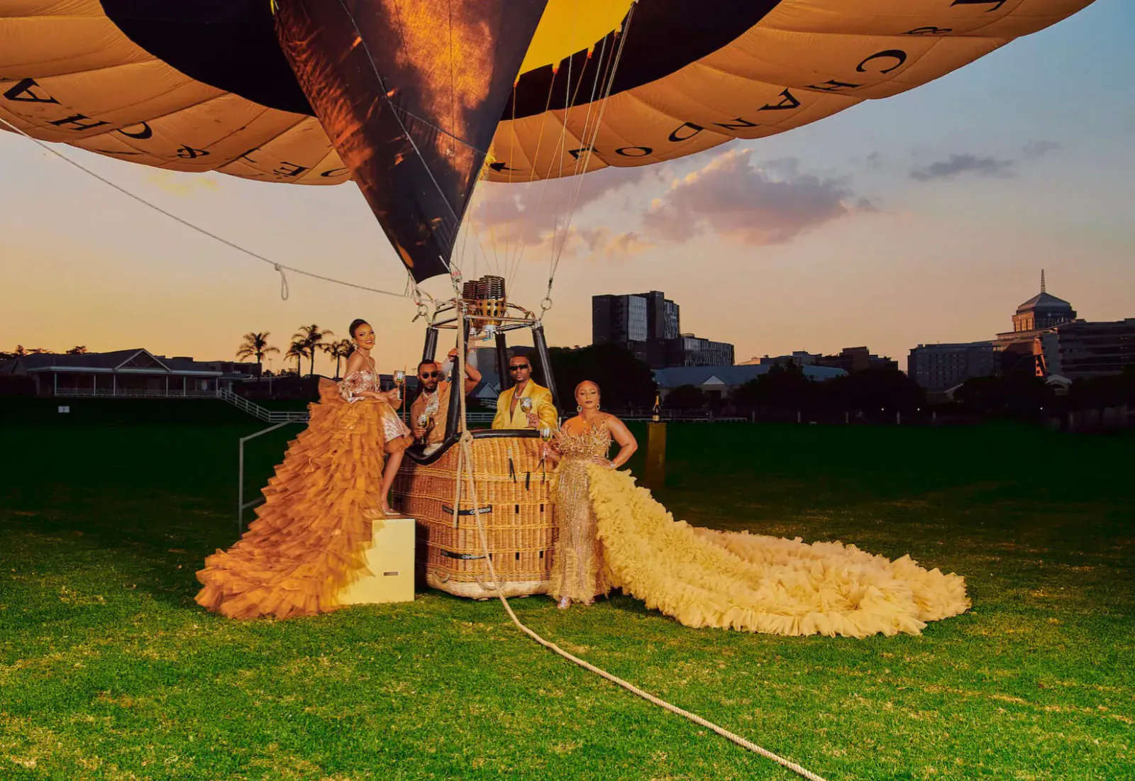 Three models in feathered gowns pose by Moët & Chandon hot air balloon basket on grass at sunset, city skyline backdrop