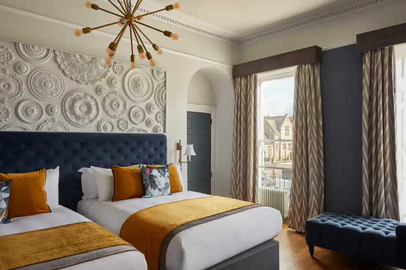 Elegant Hotel Indigo Bath bedroom with twin beds, mustard bedding, chandelier, ornate wall, large window to historic buildings.